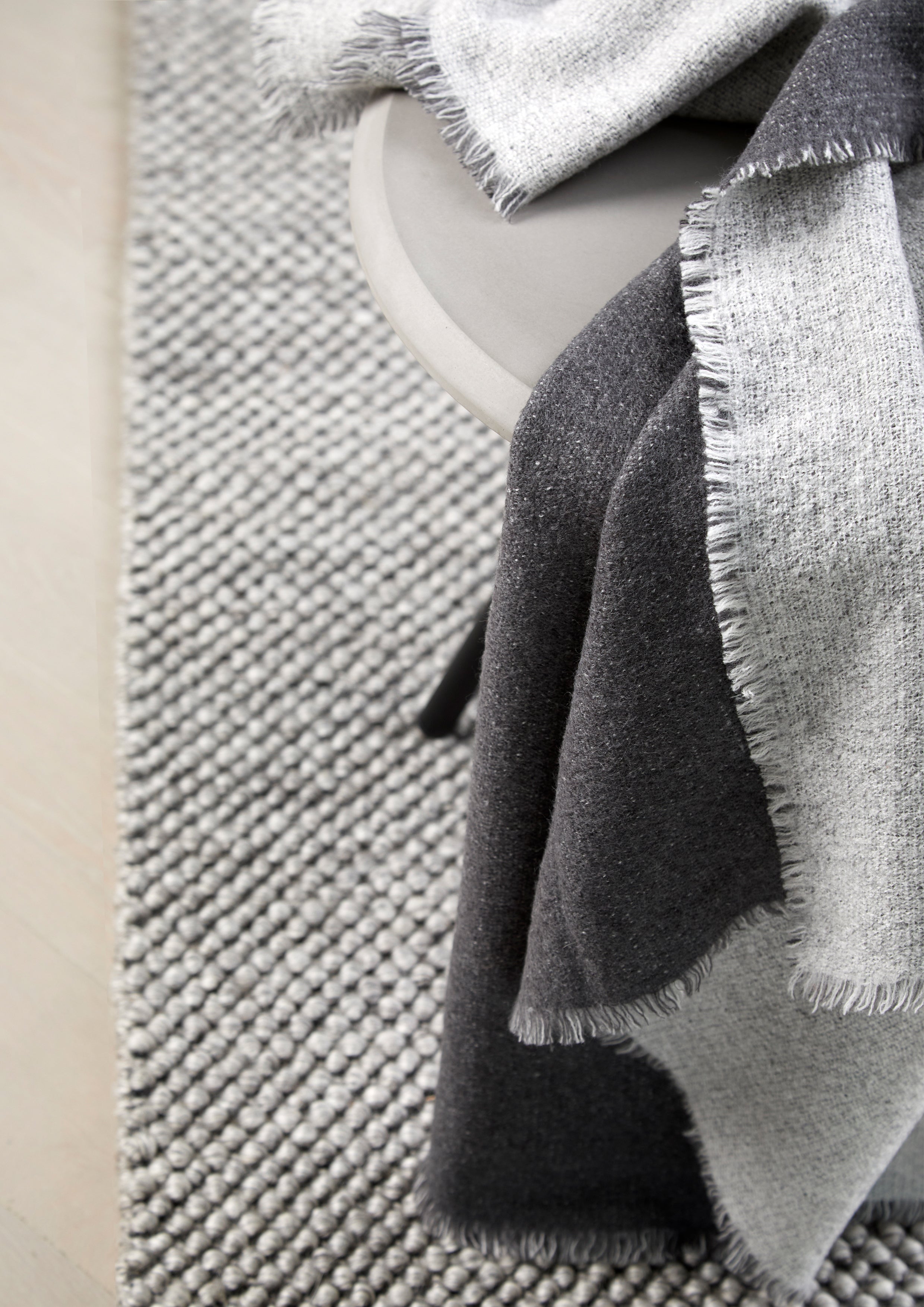 Lysegrå og mørkegrå vævet plaid i dobbeltfarvet design lagt på bord med gråt håndvævet tæppe under.