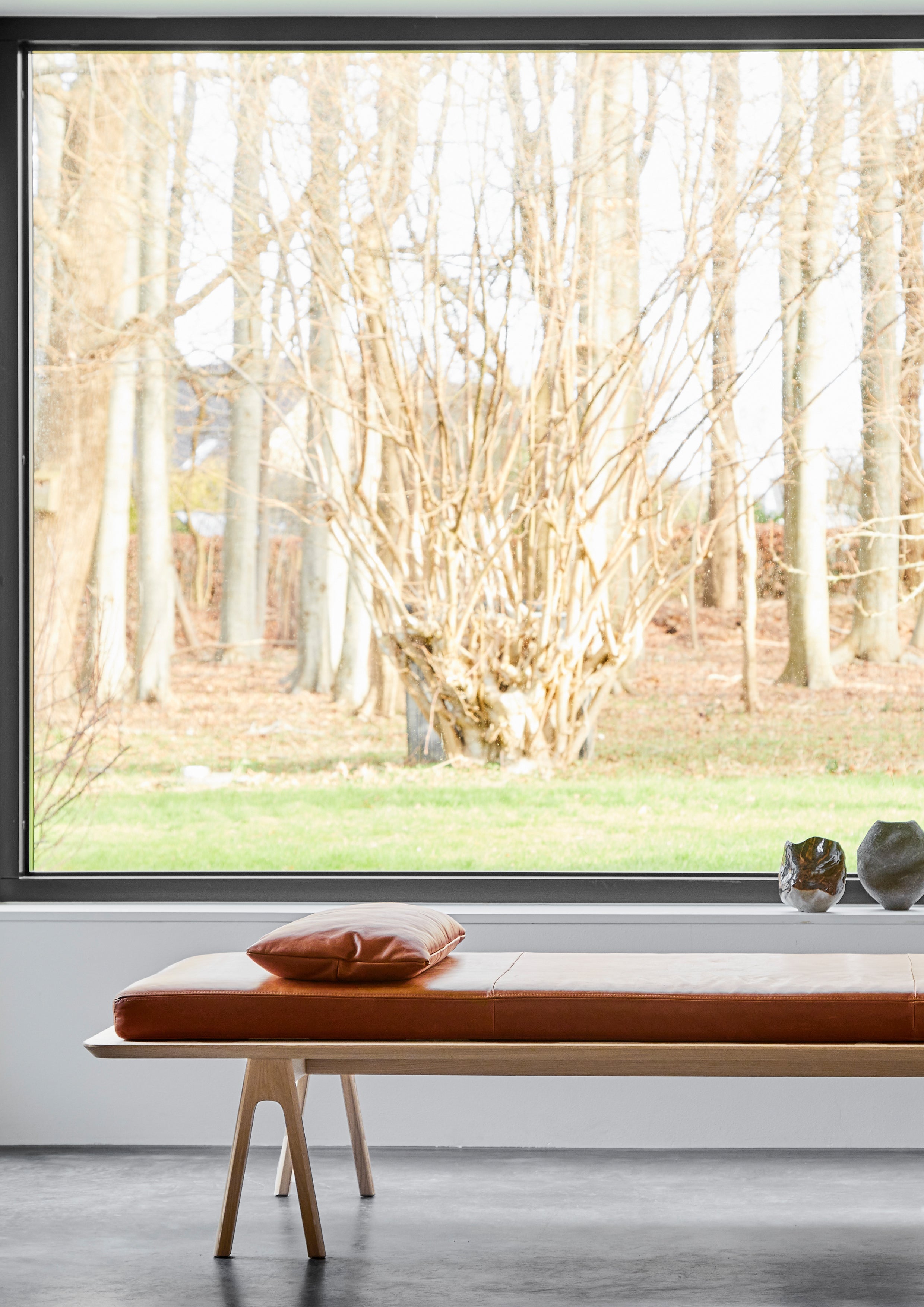 Hvidpigmenteret egetræs daybed med cognac lædermadras placeret foran vindue med udsigt til skov.