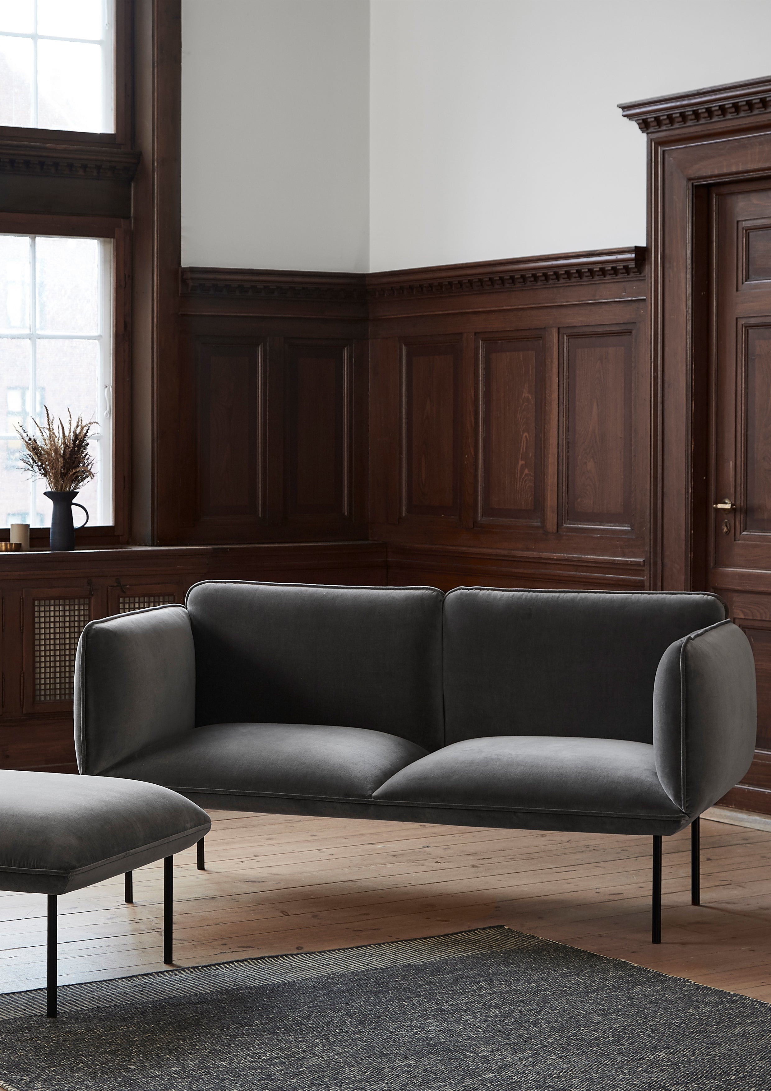 Grå velour sofa i minimalistisk design i rum med høje mørke trævægspaneler.