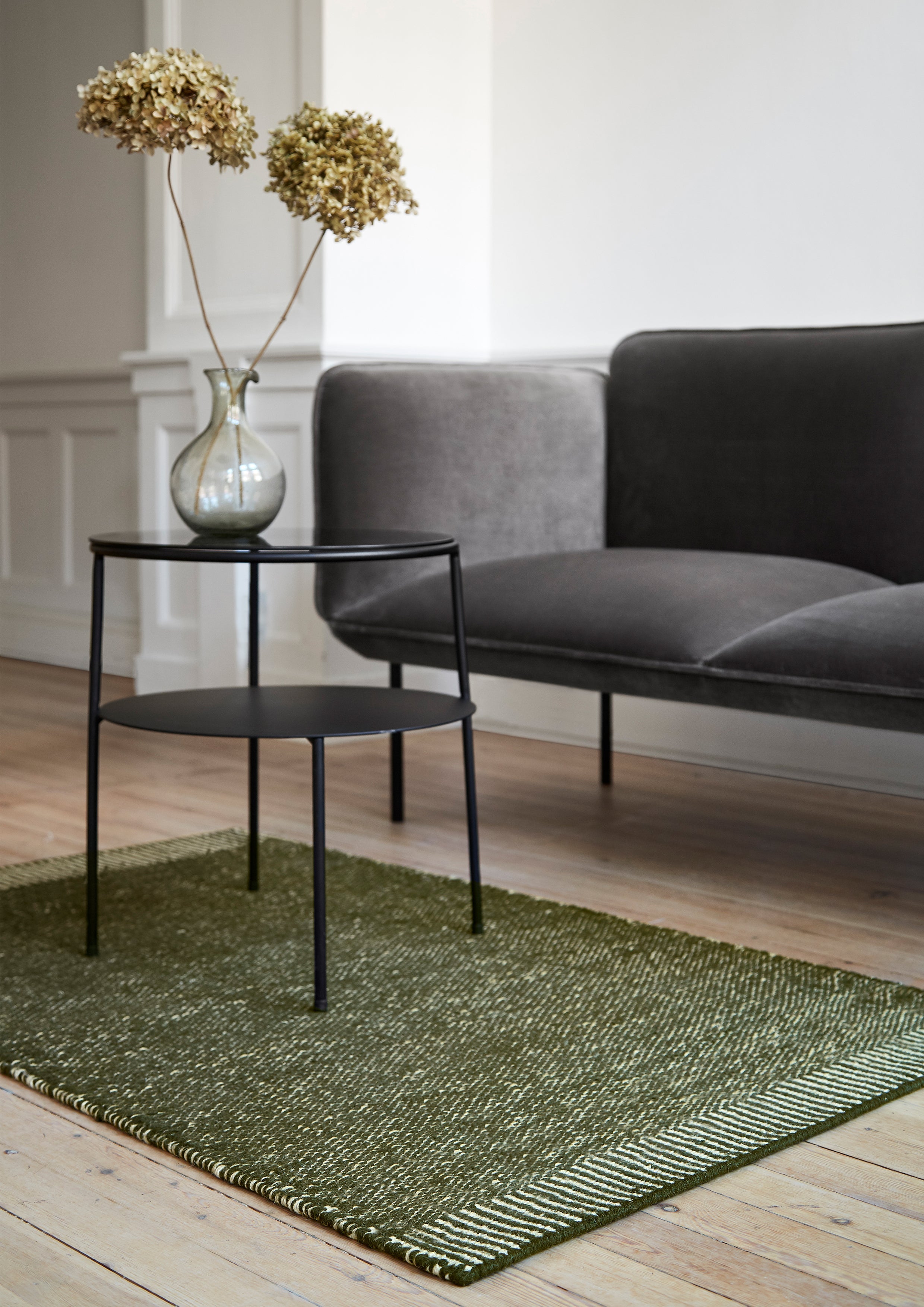 Sort sidebord foran minimalistisk sofa på grønt håndvævet tæppe.
