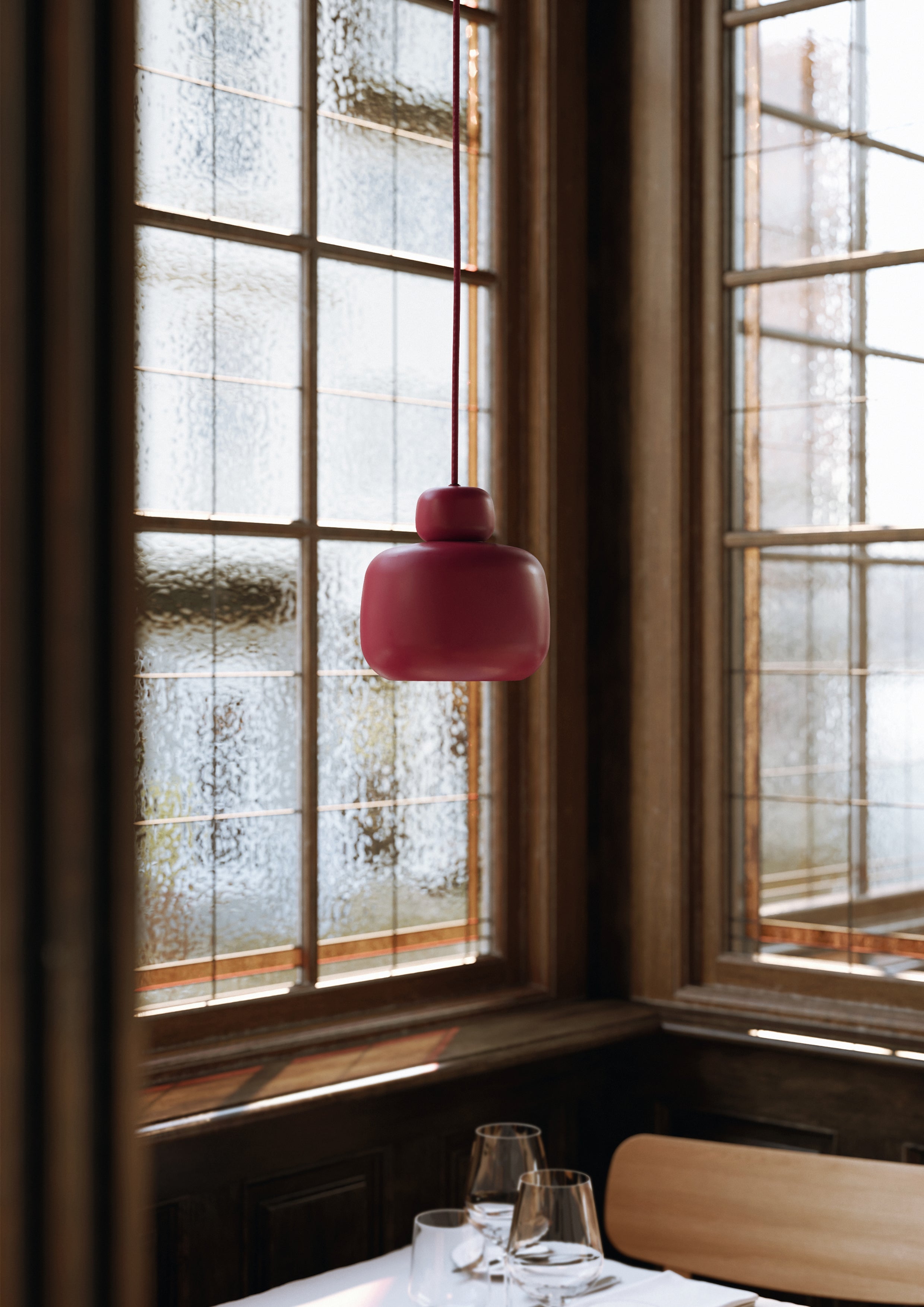 Pendel i dyb rød malet metal i skandinavisk design over restaurantbord med sprossede vinduer i baggrund.