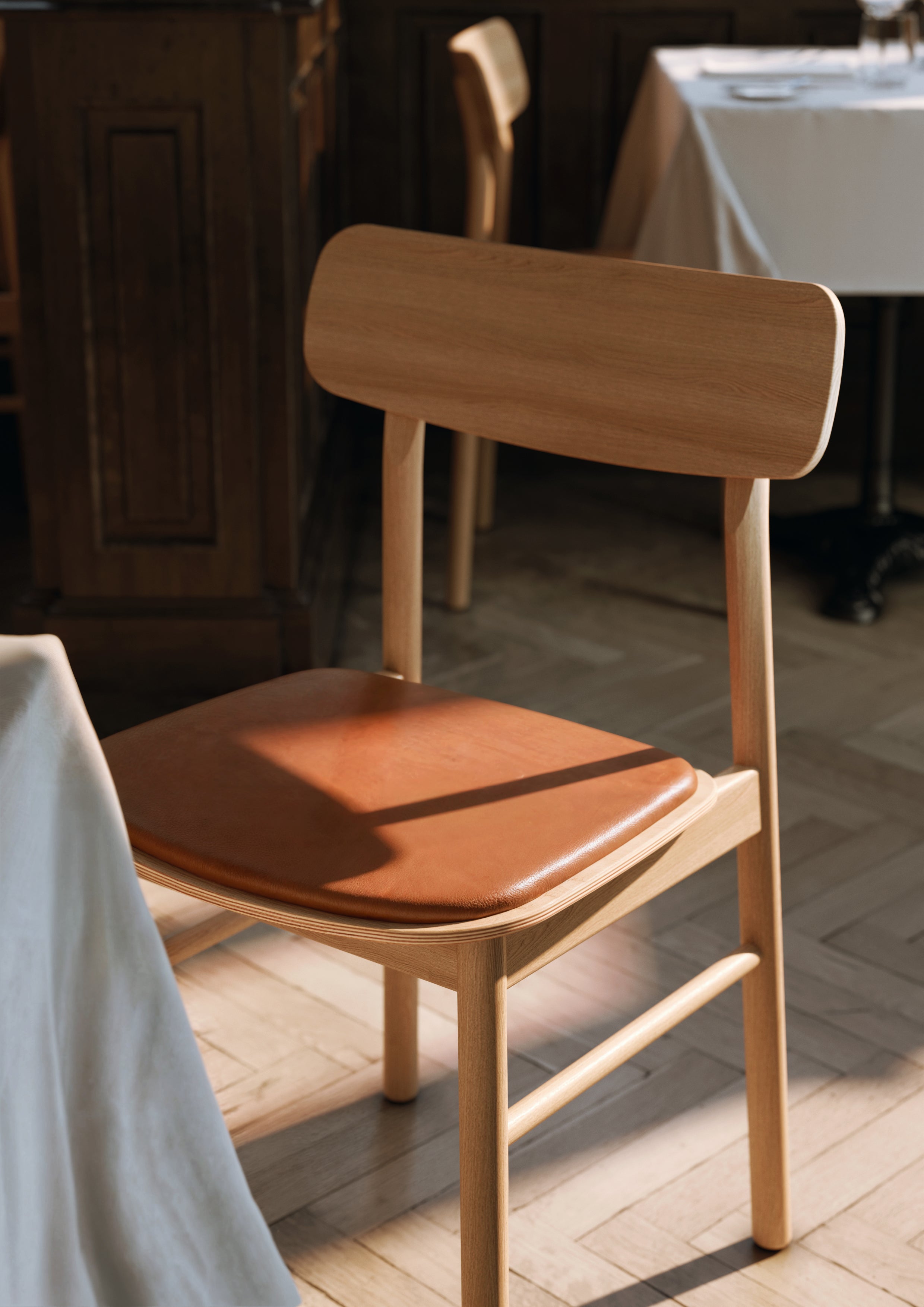 Spisebordstol i olieret egetræ med cognacfarvet lædersæde belyst af naturligt sollys i restaurant.